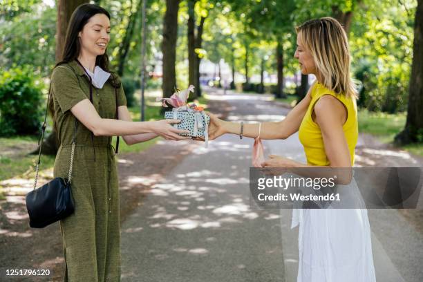 woman with taken off face mask handing over gift to friend in  nature - gift lounge stock-fotos und bilder