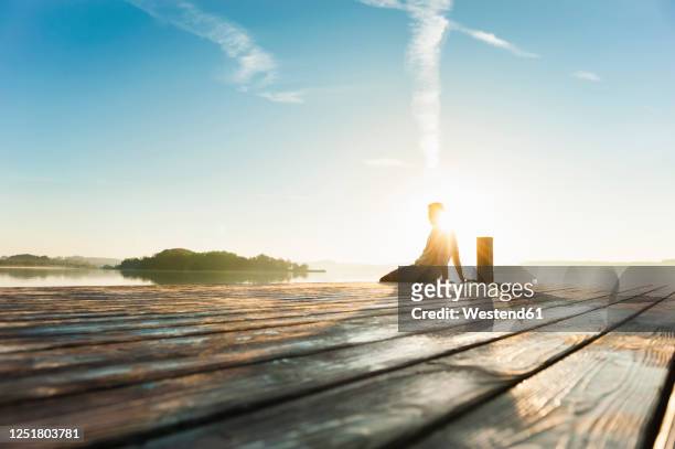young man sitting on boardwalk at lake enjoying sunrise - holzsteg stock-fotos und bilder