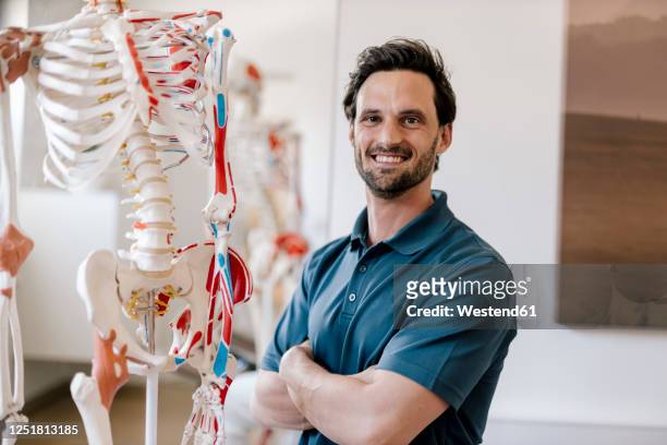 physiotherapist standing by anatomical skeleton with arms crossed - physical therapist stock pictures, royalty-free photos & images