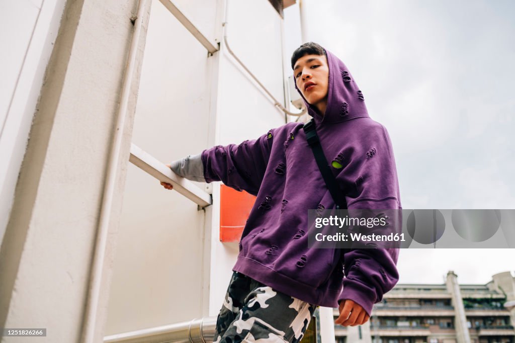 Cool young man wearing hooded shirt standing on metallic ladder against sky