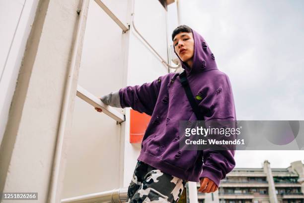 cool young man wearing hooded shirt standing on metallic ladder against sky - rebellion stock pictures, royalty-free photos & images