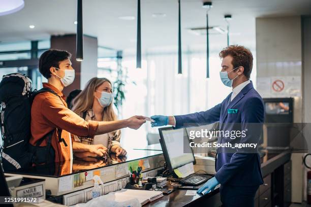 couple travelers with medical masks on hotel reception talking to male receptionist - hotel stock pictures, royalty-free photos & images