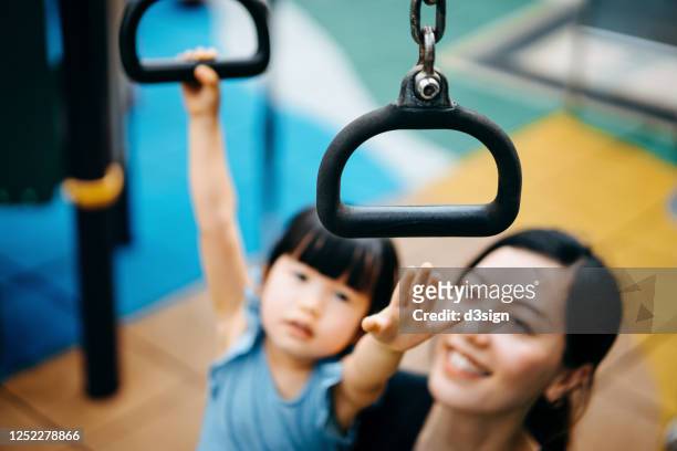 happy young asian mother supporting her little daughter while playing with the monkey bars in jungle gym. family having fun in outdoor playground on a lovely sunny day - monkey bars fotografías e imágenes de stock