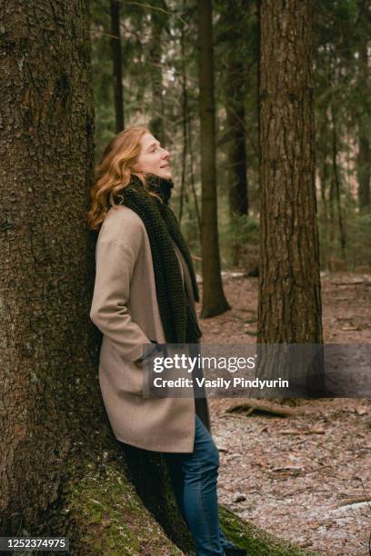 serene redhead woman in wool coat and scarf leaning against tree in woods - forest bathing stock pictures, royalty-free photos & images