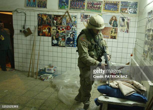 An Iraqi civilian looks at a US soldier as he searches for weapons with posters of international celebrities and soccer teams on the wall behind him,...