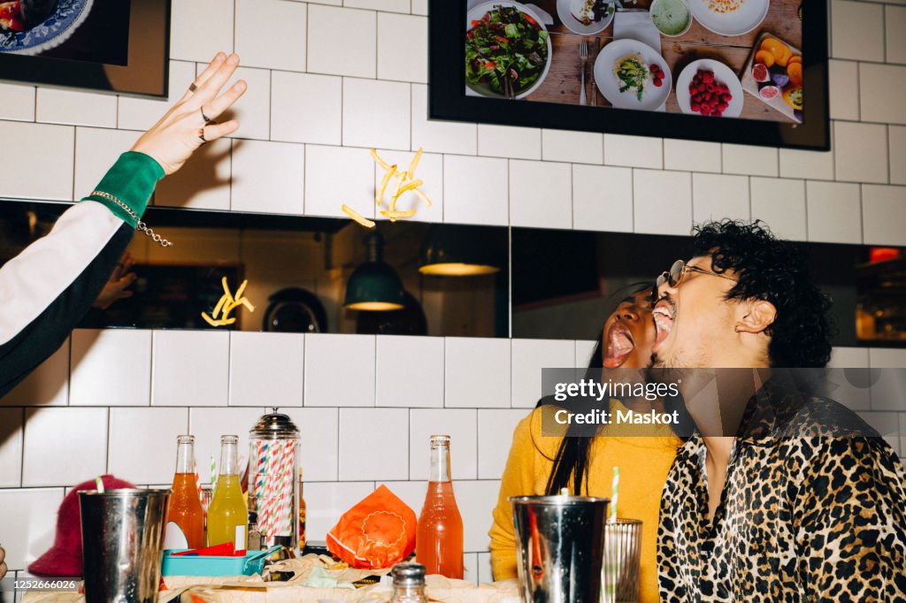 Cropped hand of man throwing french fries at male and female friends with open mouth in cafe