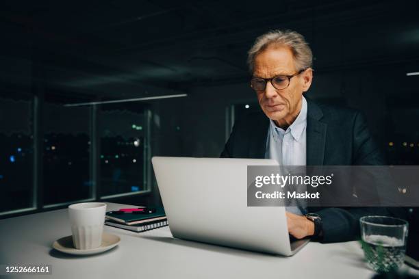 confident senior businessman using laptop while sitting at illuminated desk in creative office - male looking content stock pictures, royalty-free photos & images