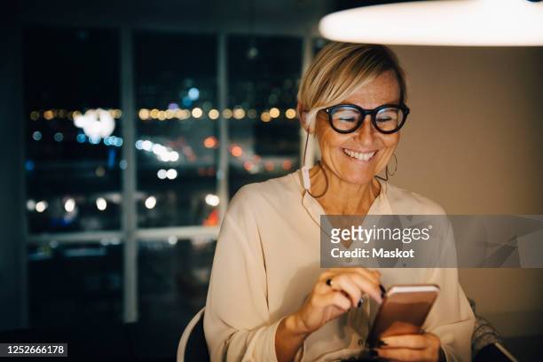 happy female entrepreneur using smart phone while sitting in illuminated office at night - medelålders kvinnor bildbanksfoton och bilder