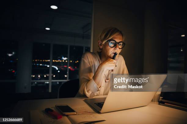 thoughtful businesswoman looking at laptop while working late in creative office - dusk dark stock-fotos und bilder