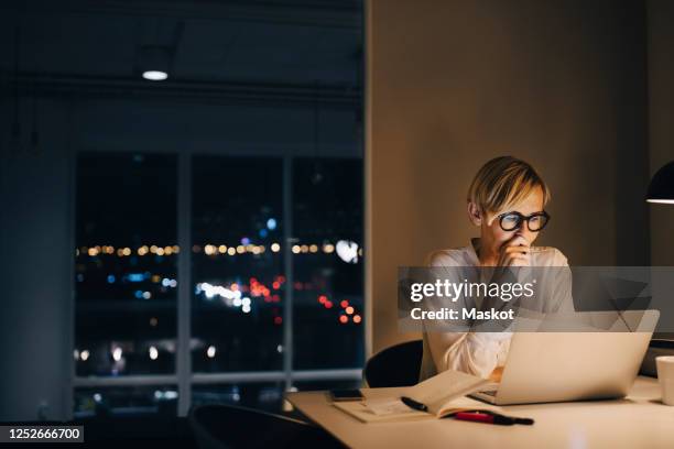 dedicated mature businesswoman using laptop while sitting at illuminated desk in coworking space - woman using smartphone with laptop stock-fotos und bilder