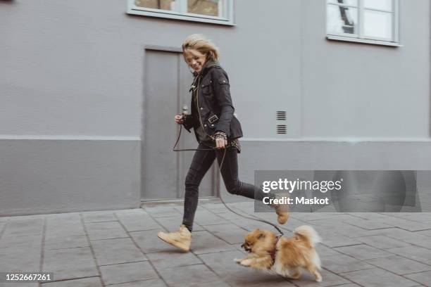 full length side view of happy woman running with her pomeranian dog on footpath by building in city - happy lady walking dog stockfoto's en -beelden