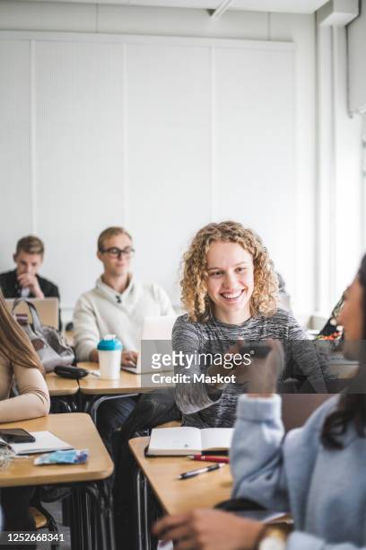 smiling young woman giving calculator to female friend while sitting in classroom - student loan stockfoto's en -beelden