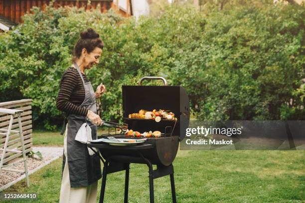 side view of smiling woman cooking dinner on barbecue grill at back yard during garden party - backyard barbeque stock pictures, royalty-free photos & images