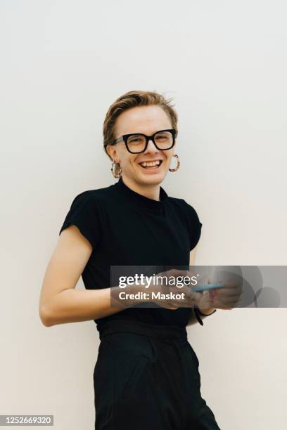 portrait of smiling androgynous businessman leaning on white wall while holding smart phone in board room - androgino fotografías e imágenes de stock