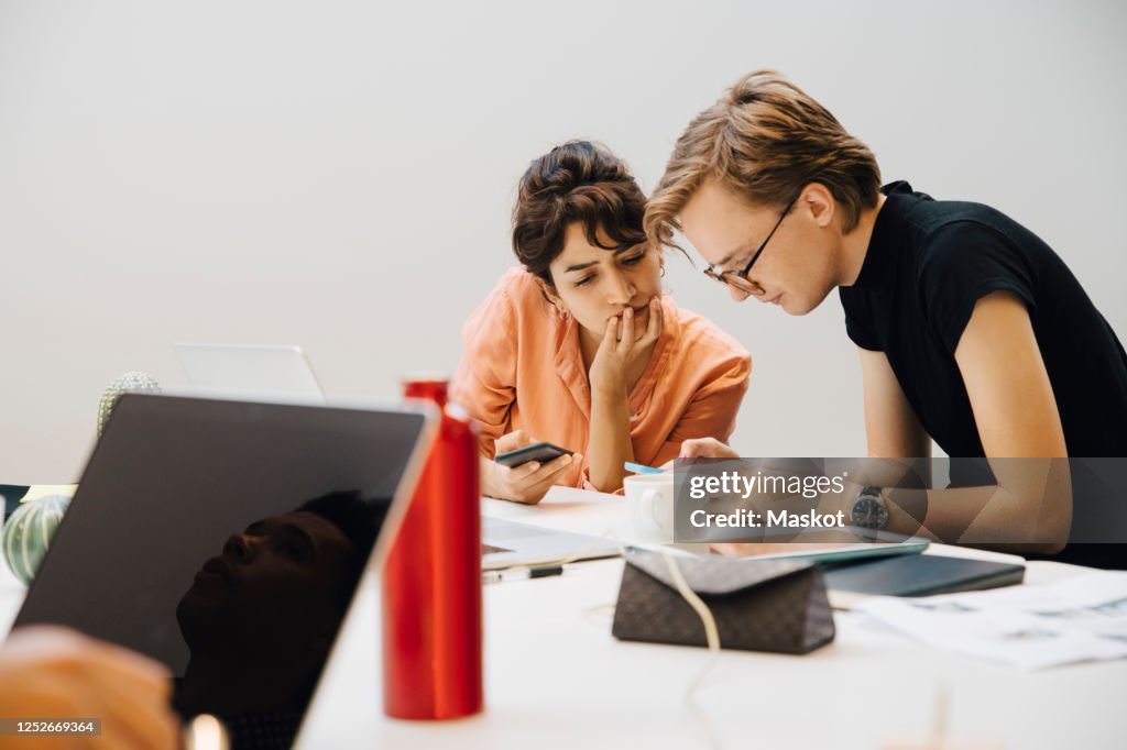 Colleagues sharing smart phones while sitting in illuminated board room during meeting