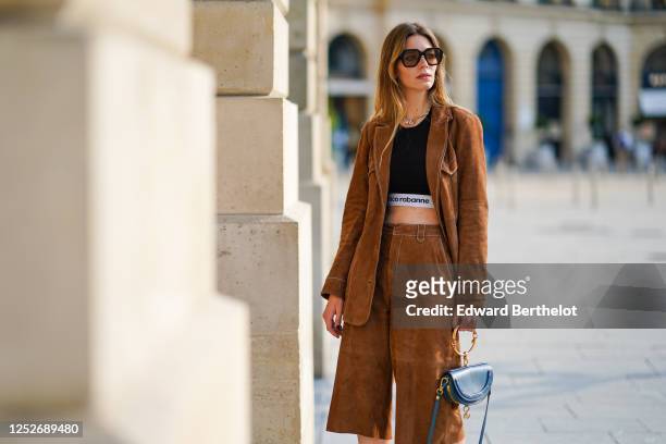 Natalia Verza wears Celine sunglasses, a necklace, a black Paco Rabanne cropped top, a Gestuz brown suede jacket, brown suede long shorts, a blue...