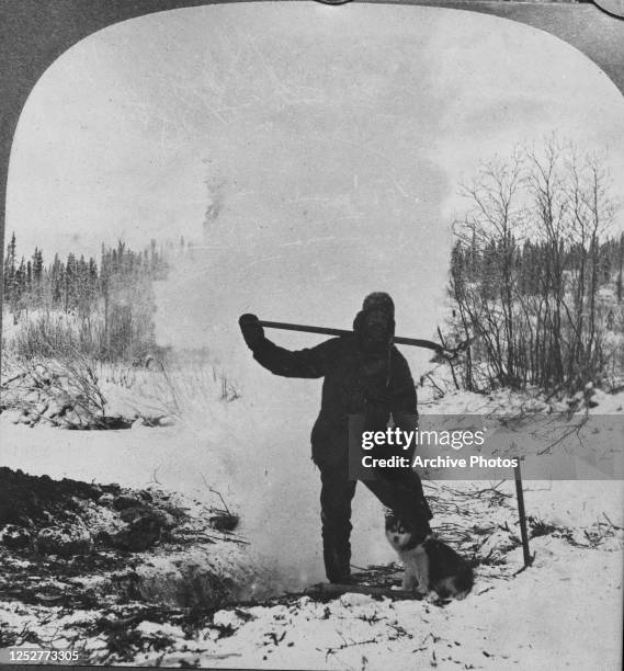 Prospector and his Alaskan Malamute puppy in Alaska during the Klondike Gold Rush, 1899. He is thawing the frozen ground with a fire in a pit.