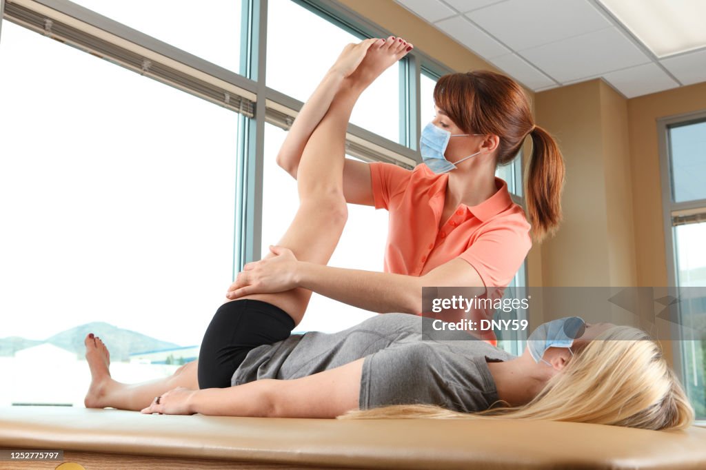 Physical Therapist Stretching The Leg Of A Female Patient While Both Wear Protective Facemasks