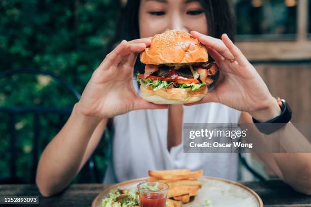 young woman eating burger at restaurant with outdoor seating - woman eating burger stock pictures, royalty-free photos & images