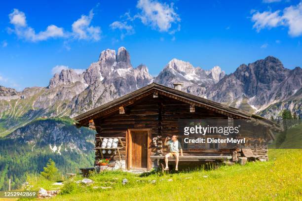 man dressed in traditional austrian or bavarian clothes sitting on bench in front of alpine hut, enjoying beer in alps - hut stock pictures, royalty-free photos & images