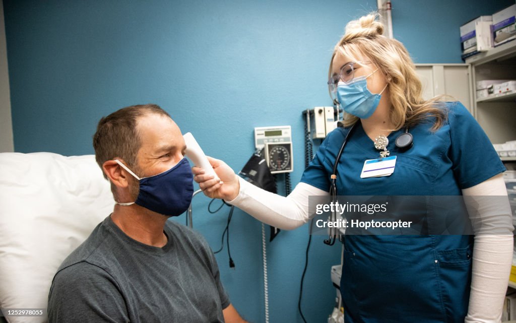 A Young Female Nurse Wearing a Surgical Face Mask Takes the Temperature of a Masked White Male in His Forties with a Forehead Infrared Thermometer in an Examination Room of a Medical Clinic