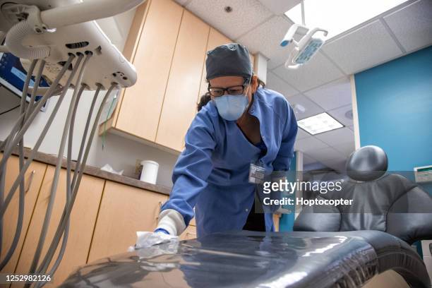 a latina healthcare worker in her fifties wearing a face mask and surgical gloves wipes a medical chair in an examination room at a dental office in preparation for the next patient - tidy room stock pictures, royalty-free photos & images