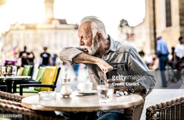 senior man at outdoor cafe coughing into elbow. - old cough stock pictures, royalty-free photos & images