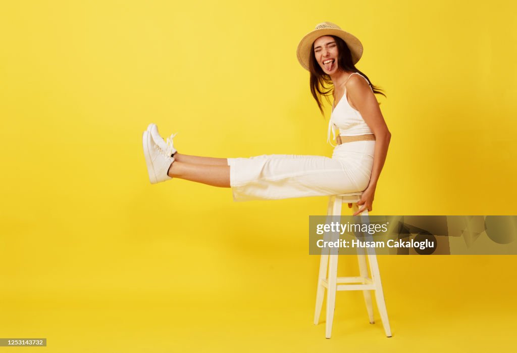 Beautiful, crazy young woman with straw hat sitting on white chair front of yellow background.
