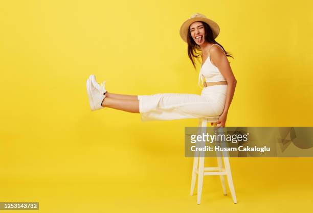 beautiful, crazy young woman with straw hat sitting on white chair front of yellow background. - legs crossed at knee stock pictures, royalty-free photos & images