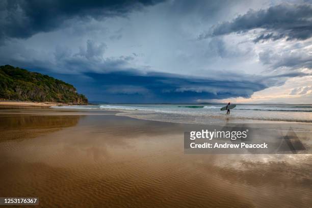 a surfer is walking along the beach at tea tree bay in noosa national park as a storm passes in the background, noosa heads, sunshine coast, queensland, australia on 9th may 2020 - noosa beach stock pictures, royalty-free photos & images