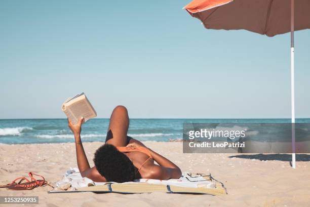 afro italian woman reading a book and relaxing on the beach in summertime - woman on beach reading stock pictures, royalty-free photos & images