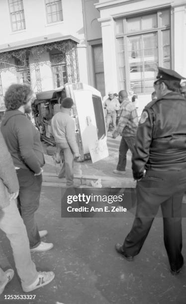 View of bystanders, including a police officer, gathered near a Chevrolet station wagon, tipped on its side during an anti-KKK demonstration, in...