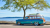 Vintage car parked on the tropical beach (seaside) with surfboard on the roof. Leisure trip in the summer. Retro color effect. West coast Pacific ocean.