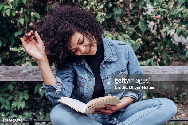 adorable young african american woman reading a good book while chilling on bench in park - reading stock-fotos und bilder