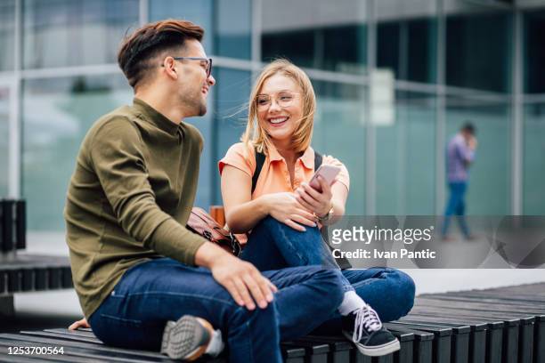 zwei lächelnde studenten sitzen auf einer parkbank - student stock-fotos und bilder