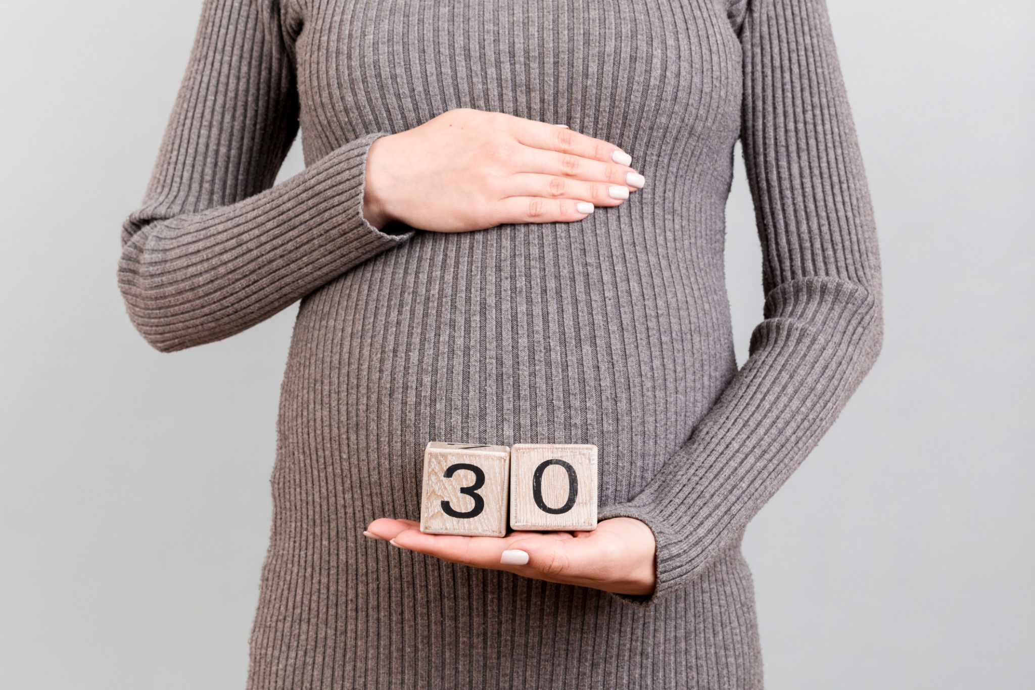 Close up of pregnant woman in gray dress holding cubes with numbers of pregnancy weeks. Thirty weeks of pregnancy period at gray background. Copy space Close up of pregnant woman in gray dress holding cubes with numbers of pregnancy weeks. Thirty weeks of pregnancy period at gray background. Copy space