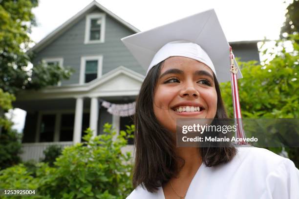 A teenage girl on her graduation day outside in front of a house in her neighborhood. She is wearing a white cap and gown. She is smiling and very happy.