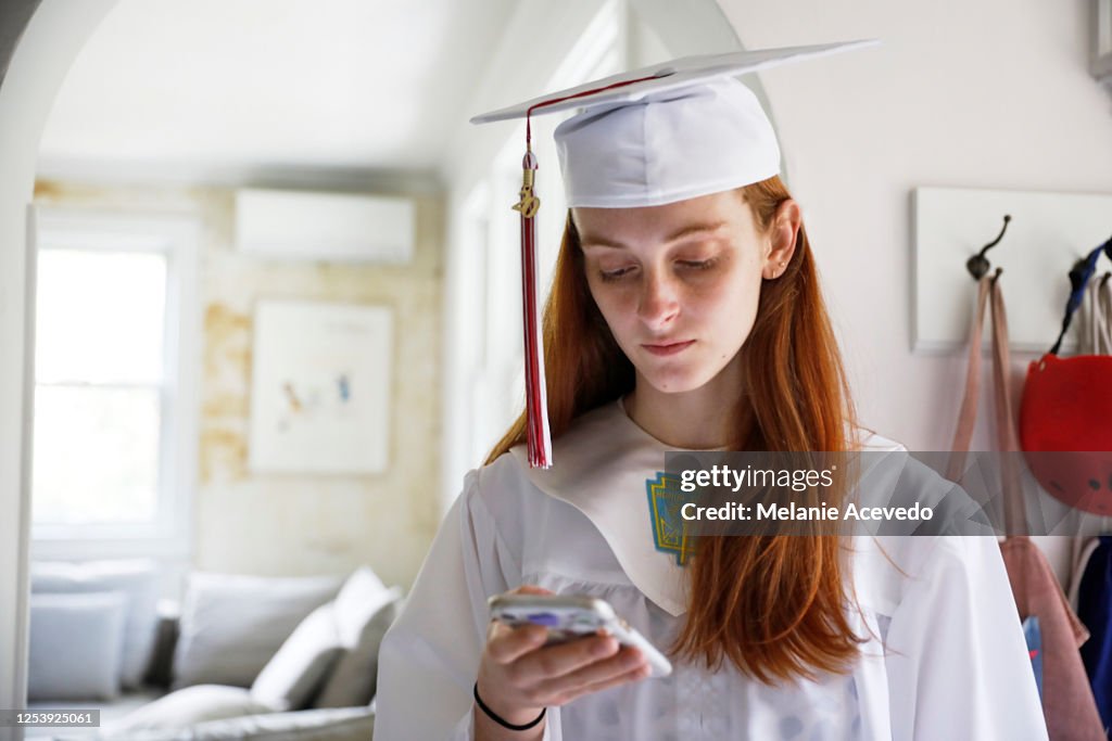 Redheaded teenage girl wearing a cap with red tassel and white graduation gown on graduation day looking down at her phone inside her home preparing for graduation ceremony