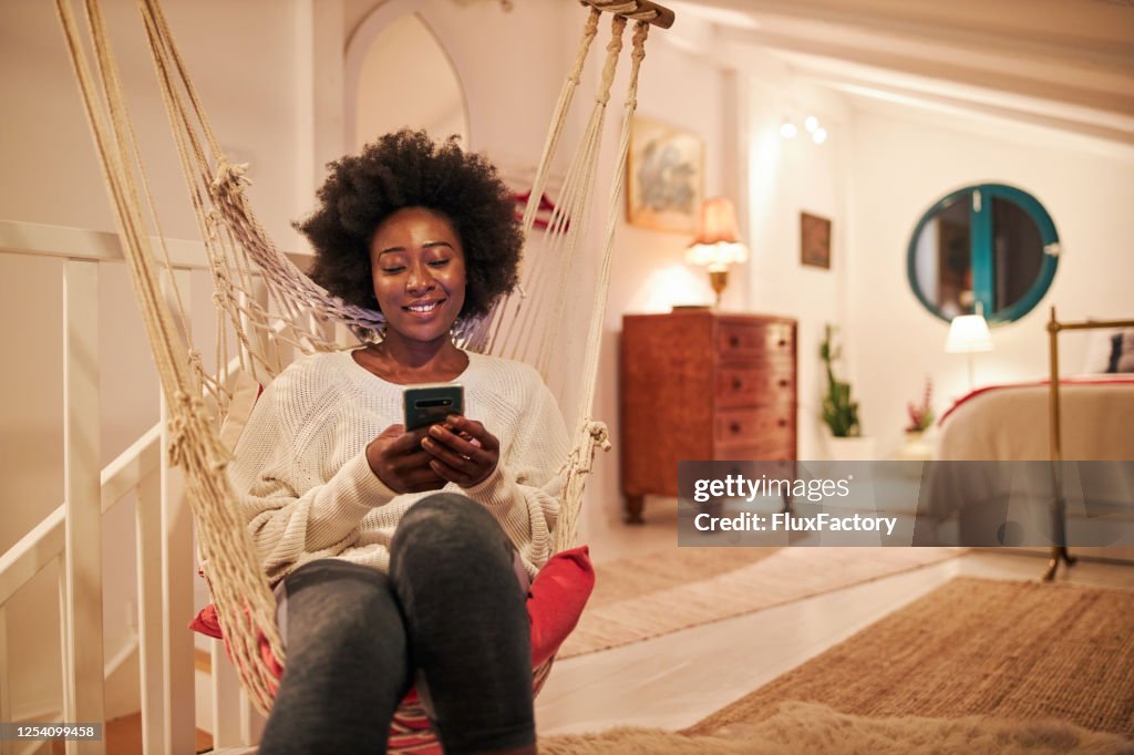Cheerful young woman using a mobile phone while sitting in a swing at home