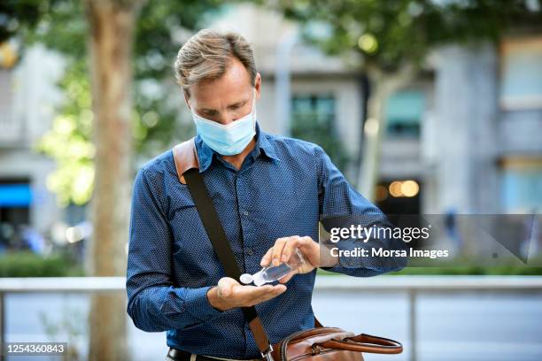 businessman disinfecting hands with hand sanitizer during pandemic in city. he is wearing protective face mask. - hand sanitiser - fotografias e filmes do acervo