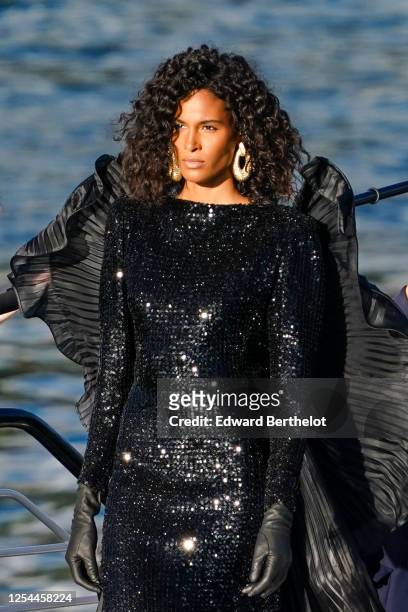 Cindy Bruna wears golden large earrings, a black glittering sequined shiny dress, a ruffled pleated cape, black leather gloves, during the "Balmain...