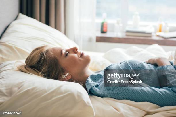 glimlachende vrouw die in bed ligt en aan muziek door haar draadloze oortelefoons luistert - luisteren stockfoto's en -beelden