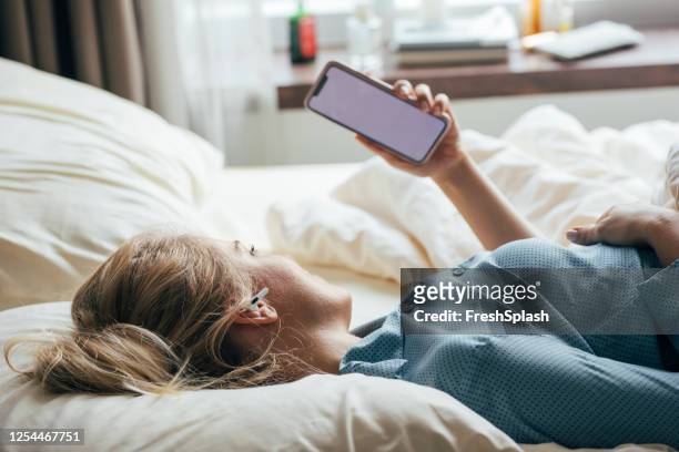 blonde woman in pajamas lying in bed and using her smartphone to watch something - read and newspaper and bed stock pictures, royalty-free photos & images