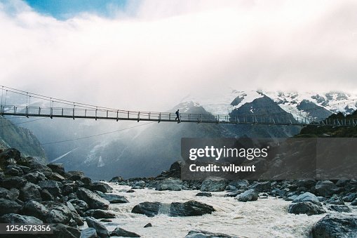 Female Hiker crossing a suspension bridge over a glacier river with snowcapped mountains and a glacier behind Female Hiker crossing a suspension bridge over a glacier river with snowcapped mountains and a glacier behind