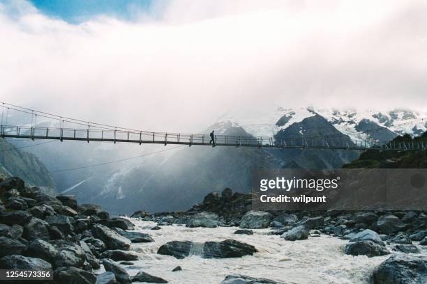 female hiker crossing a suspension bridge over a glacier river with snowcapped mountains and a glacier behind - nepal stock pictures, royalty-free photos & images
