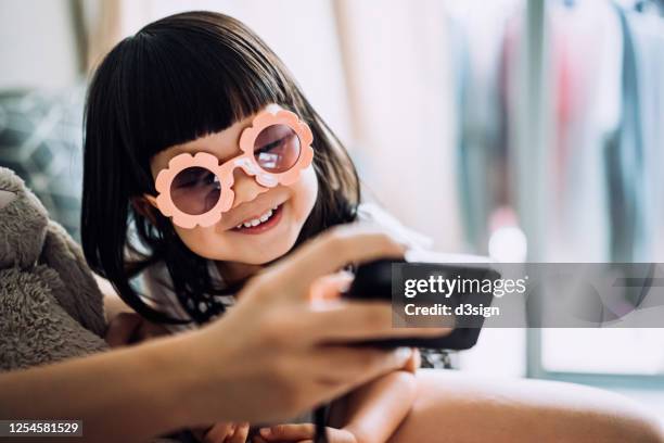 cute little asian girl with flower-shaped sunglasses and her mother smiling joyfully while looking at the photos taken on camera in the living room at home - child playing in room stockfoto's en -beelden