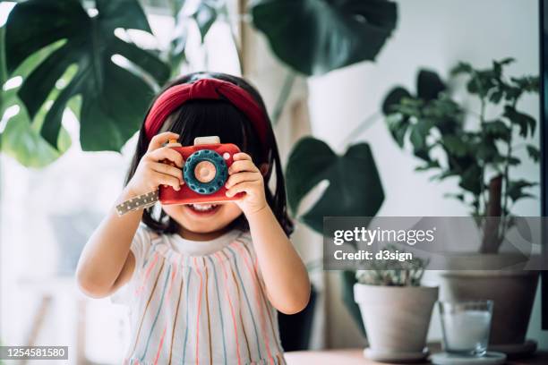 smiling little asian girl with red headband acting like a professional photographer having fun while taking photos with wooden toy camera in front of potted plants at home - creative play stock-fotos und bilder