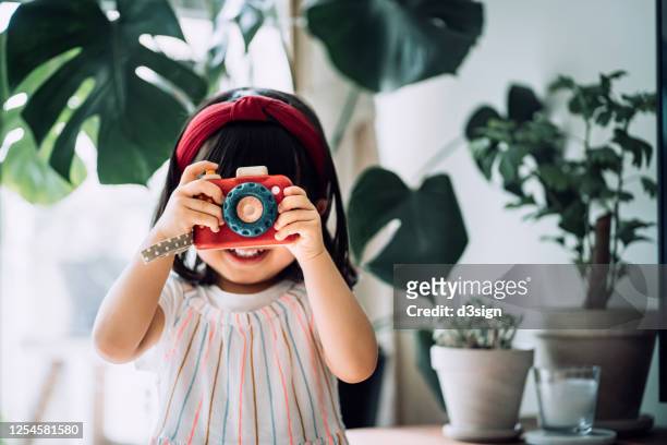 smiling little asian girl with red headband acting like a professional photographer having fun while taking photos with wooden toy camera in front of potted plants at home - children camera stock pictures, royalty-free photos & images