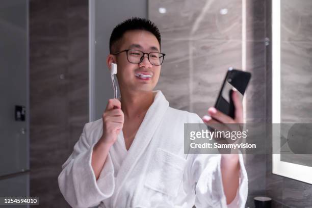 asian man brushing teeth in bathroom while using phone - brush teeth phone stock-fotos und bilder