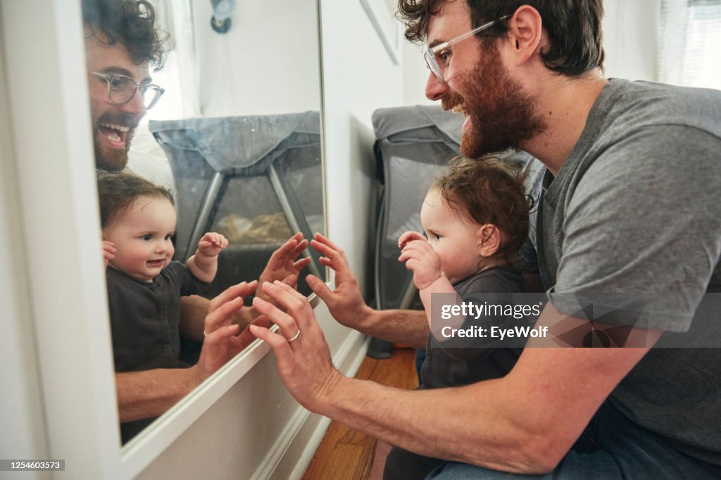 A father sitting in a bedroom with his baby daughter looking into a mirror, smiling.